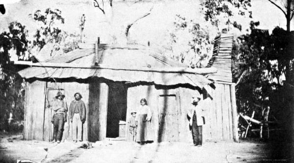 William Thomas Guardian of the aboriginals in Westernport and Mordialloc Districts, outside a colnial hut with three adult aboriginals and one child [picture].